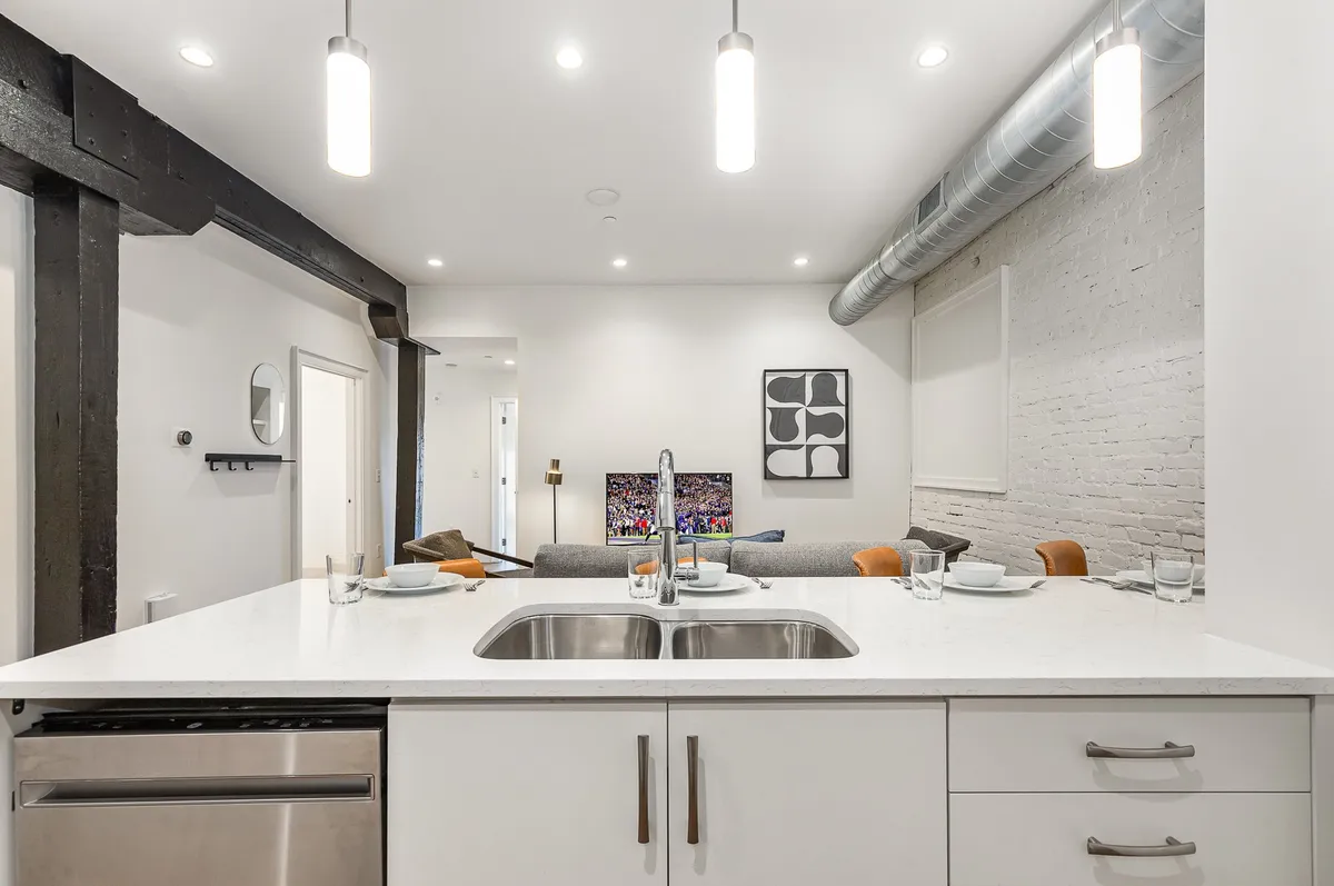 An open-concept kitchen and living area featuring a kitchen island with a stainless steel sink, modern lighting fixtures, nearby sofa, and artwork. The setup encourages conversation and interaction between guests while cooking or dining.