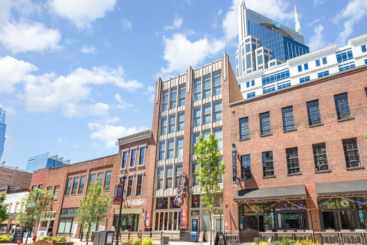 The image showcases a vibrant Nashville street scene just outside the property, with historic brick buildings juxtaposed with a sleek modern skyscraper. Lush greenery and outdoor seating areas invite guests to linger and soak up the energetic atmosphere. People are enjoying outdoor cafes, exploring local shops, and gathering for cultural events in a lively, artistic square.