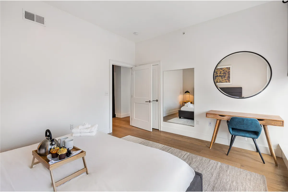 A tranquil bedroom showcasing a cozy bed topped with a breakfast tray, a desk and chair setup ideal for catching up on work, and a mirror that enhances the room's airy feel. The space is styled for both comfort and practicality, representing restful nights and functional amenities.