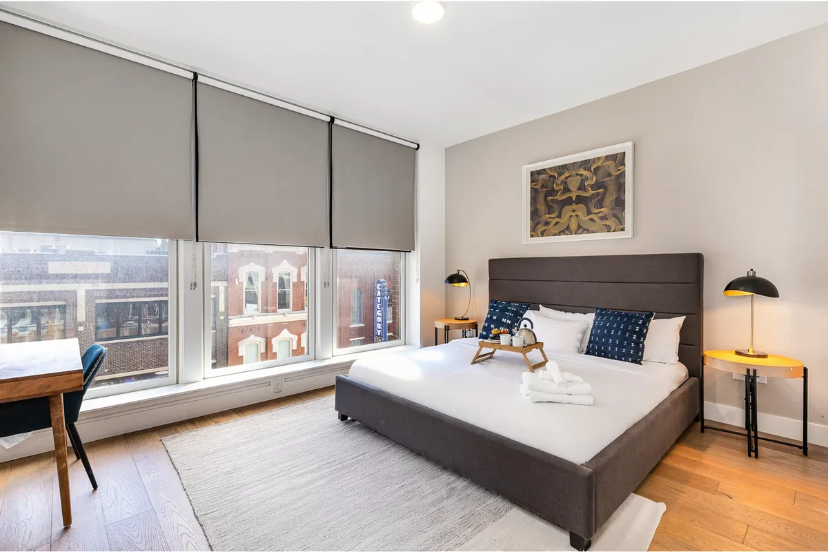 A stylish bedroom suite featuring a bed with crisp white linens, large windows offering city views, contemporary artwork, and a wooden table with a chair that doubles as a workstation. Natural light fills the room, creating a restful and inviting space ideal for both relaxation and productivity.
