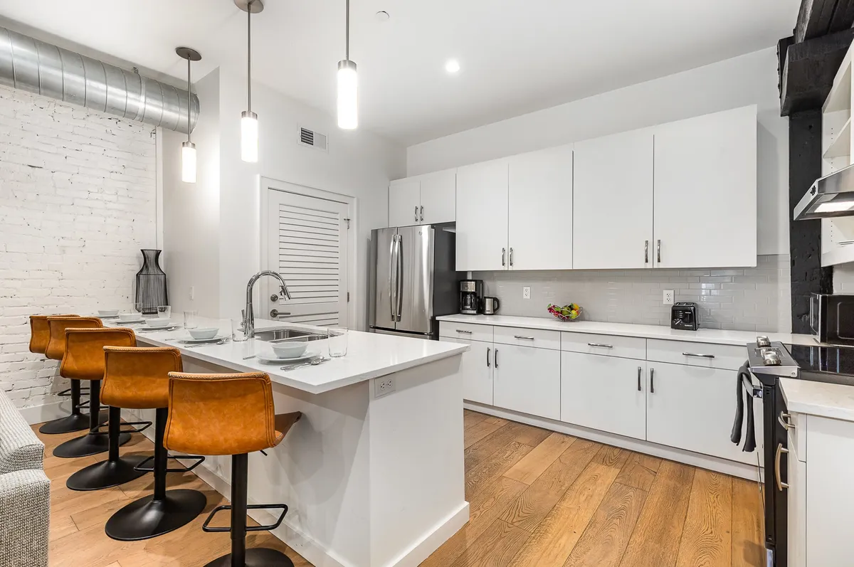 This image features a bright, contemporary kitchen with a large island at its center, surrounded by bar stools. White cabinetry, stainless steel appliances, and an exposed brick wall create a modern yet inviting atmosphere. The island offers ample space for dining or food prep, perfectly suited for group gatherings.